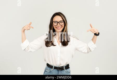 Cheerful brunette woman with glasses pointing to herself with both thumbs, wearing a white shirt Stock Photo
