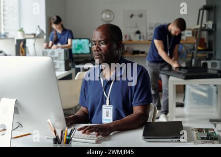Serious mature male worker of repair service center touching keys of desktop computer keyboard and looking at screen by workplace Stock Photo