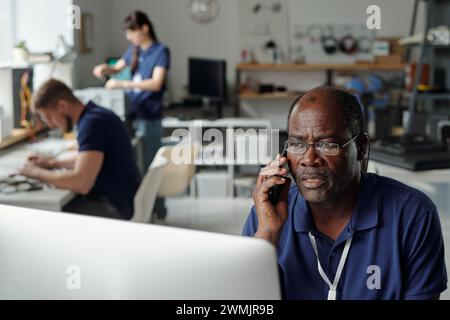 Mature male technician of repair service sitting by workplace in front of computer screen and talking to client on mobile phone Stock Photo