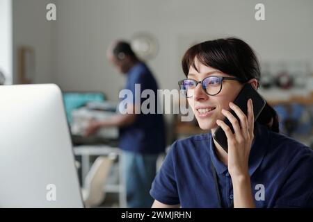Smiling female technician with mobile phone talking to client while sitting in front of computer screen and looking through online data Stock Photo