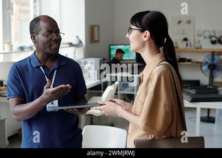 Mature male technician passing laptop to young smiling female client after checking it and identifying technical problems during consultation Stock Photo