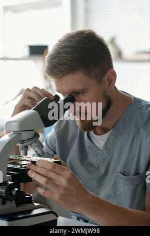 Young serious male technician of troubleshooting center looking at motherboard in microscope while sitting by workplace Stock Photo