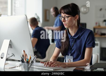 Young female worker of repair service center typing on computer keyboard and looking at screen while entering data in coworking space Stock Photo