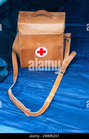 Vintage survival. Brown leather first aid bag with strap and red cross on white round shape. Inside blue scout tent. Stock Photo