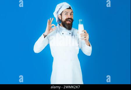 Handsome bearded chef man in uniform holds bottle with milk. Happy male cook in apron with bottle milkshake or yogurt showing sign ok. Drinks, dairy Stock Photo