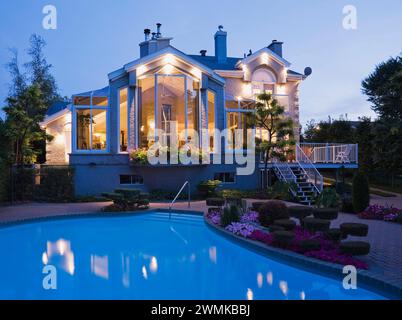 Modern home with large glass panoramic windows and landscaped backyard with in-ground swimming pool at dusk. Stock Photo