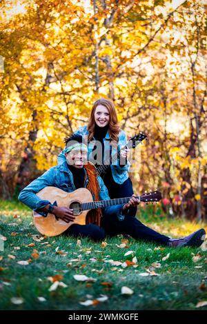 Portrait of a mixed race married couple, smiling and posing for the camera holding guitars while sitting on the grass during a fall family outing i... Stock Photo