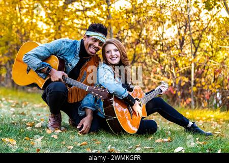 Portrait of a mixed race married couple, smiling and posing for the camera holding guitars while sitting on the grass during a fall family outing i... Stock Photo