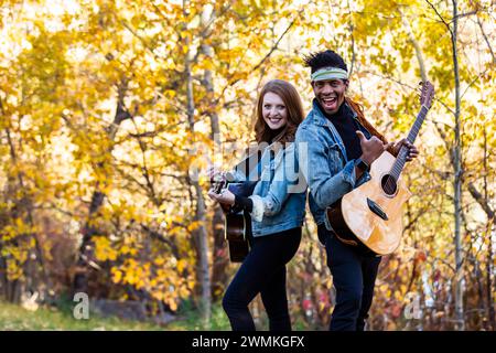 Mixed race married couple standing back to back, smiling and posing for the camera playing guitars during a fall family outing in a city park, spen... Stock Photo