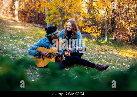 View through leaves of a mixed race married couple sitting on grass, spending quality time together, smiling and playing guitars during a fall fami... Stock Photo