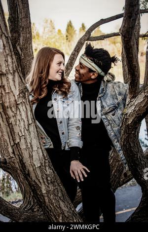 Close-up portrait of a mixed race couple smiling at each other while resting on tree branches, spending quality time together during a fall family ... Stock Photo