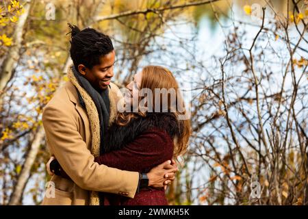 A mixed race couple standing by the shore of a lake hugging each other while spending quality time together during a fall family outing in a city park Stock Photo