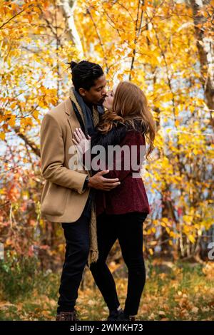 A mixed race couple standing by the shore of a lake hugging and kissing each other while spending quality time together during a fall family outing... Stock Photo