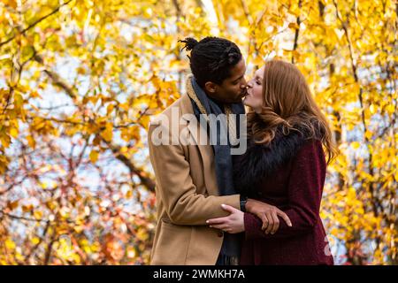 A mixed race married couple hugging and kissing each other while spending quality time together during a fall family outing in a city park Stock Photo