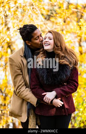 A mixed race married couple hugging with husband kissing wife on the cheek while spending quality time together during a fall family outing in a ci... Stock Photo