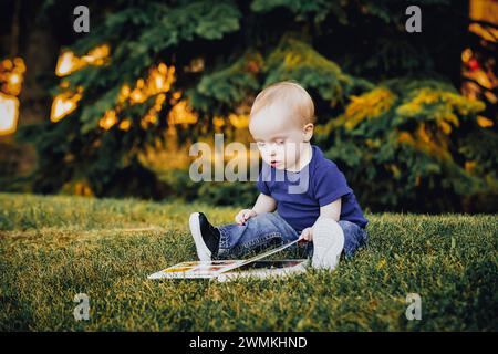 Baby boy with Down syndrome sitting on grass in a city park and looking at a book during a warm fall afternoon; Leduc, Alberta, Canada Stock Photo