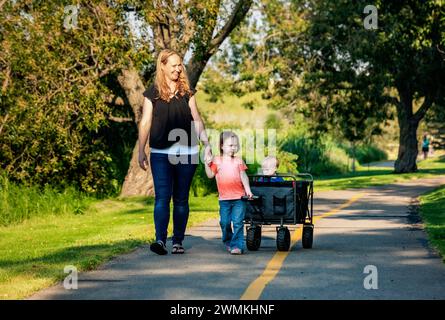 Mother and her preschooler daughter pulling a wagon with her young son who has Down Syndrome in a city park during an autumn day Stock Photo