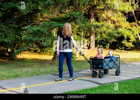 Mother pulling a wagon with her young son who has Down Syndrome and her preschooler daughter, in a city park during an autumn day Stock Photo