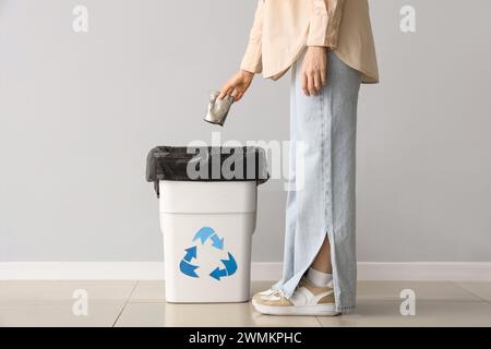 Woman throwing tin can into trash bin with recycle logo near grey wall Stock Photo