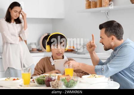 Angry father taking tablet computer away from his teenage son at table during dinner in kitchen. Family problem concept Stock Photo