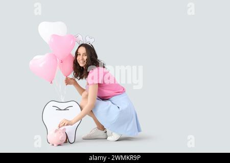 Tooth Fairy with balloons putting coin into piggy bank on light background Stock Photo