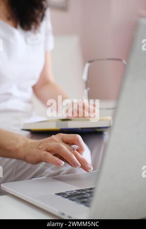 A medical worker is focused on typing on a laptop keyboard while holding a notebook on their lap, possibly recording patient information or scheduling Stock Photo