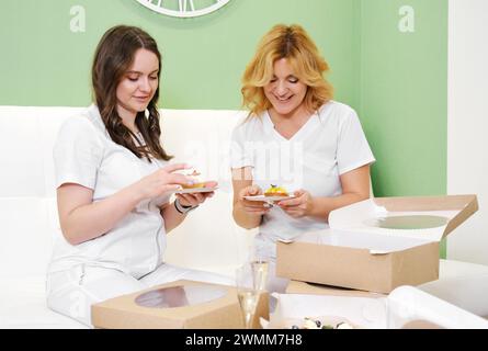 Two cheerful healthcare workers in white attire are having cupcakes while taking a break in a bright room with green walls. Horizontal photo Stock Photo