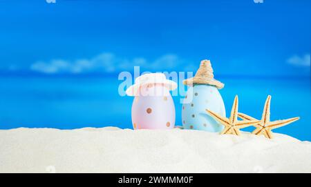 Pink and blue with polka dot eggs with hat with starfish on beach with sea on sunny day. Easter, travel, vacation. Copy space Stock Photo