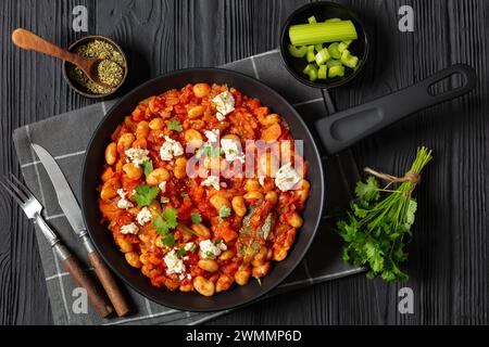 gigantes plaki, greek giant baked beans in a chunky tomato sauce sprinkled with feta cheese and fresh cilantro on skillet on black wooden table with r Stock Photo