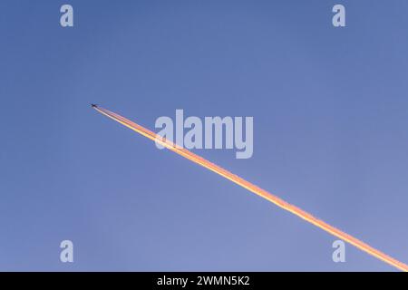 A Boeing 747 jumbo jet flying high in a cloudless blue evening sky, with its long straight contrail lit pink by the setting sun (UK) Stock Photo