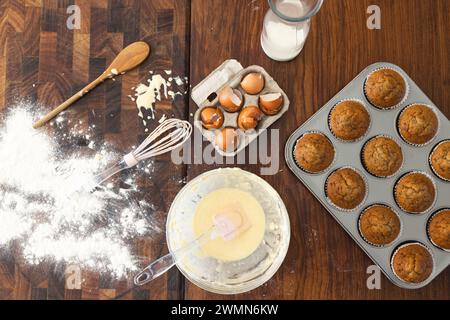 Baking ingredients and utensils are spread out on a kitchen table. Freshly baked muffins cool next to a bowl of batter, signaling a baking session at Stock Photo