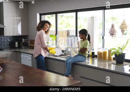 Biracial mother and daughter share a moment in the kitchen at home. They enjoy a casual conversation while doing household chores together. Stock Photo