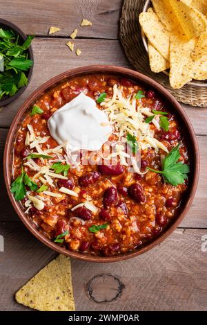 Chili con carne with cheese topings and nachos on wooden table, top view. Homemade mexican food, chili with red beans and ground beef. Chilean beans. Stock Photo