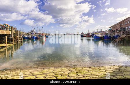 Fishing boats are moored on two sides of a harbour with a marina in the background. The sea laps over a cobbled slipway and a sky with cloud is above. Stock Photo