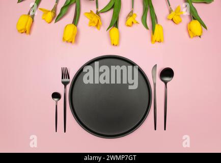Top view of yellow tulips and daffodils on pink background. Table setting, black plate and cutlery. Spring colourful composition, flat lay, copy space Stock Photo