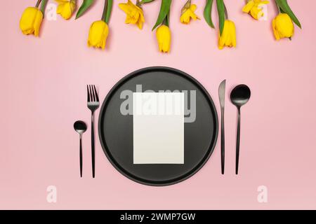 Top view of yellow tulips and daffodils on pink background. Table setting, black plate and cutlery, white card. Spring colourful composition, flat lay Stock Photo
