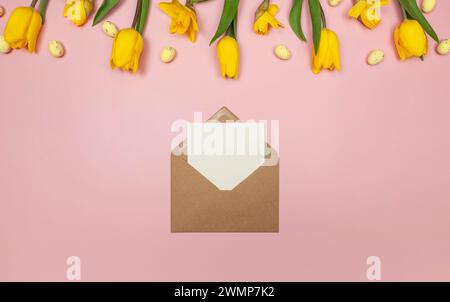 Top view of yellow tulips, daffodils, easter eggs on pink background. Kraft envelope, white card. Spring easter composition, flat lay, copy space. Stock Photo