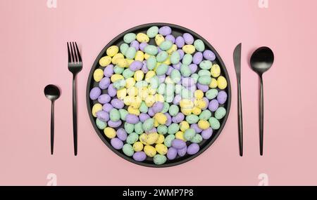 Top view of colourful easter eggs in black plate on pink background. Table setting, black plate and cutlery. Creative easter composition, spring. Stock Photo
