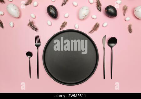 Top view of easter eggs, feathers on pink background. Table setting, black plate and cutlery. Creative easter composition, spring, flat lay. Stock Photo