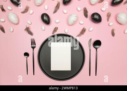 Top view of easter eggs, feathers on pink background. Table setting, black plate and cutlery, white card. Creative easter composition, spring. Stock Photo
