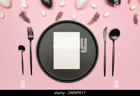 Top view of easter eggs, feathers on pink background. Table setting, black plate and cutlery, white card. Creative easter composition, spring. Stock Photo