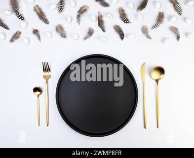 Top view of brown feathers, white easter eggs on white background. Table setting, black plate and gold cutlery. Creative easter composition, spring. Stock Photo