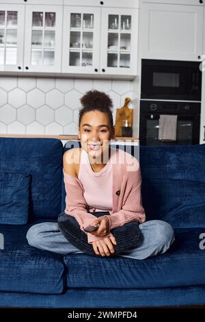 good looking jolly african american woman in homewear sitting on sofa with remote and watching TV Stock Photo