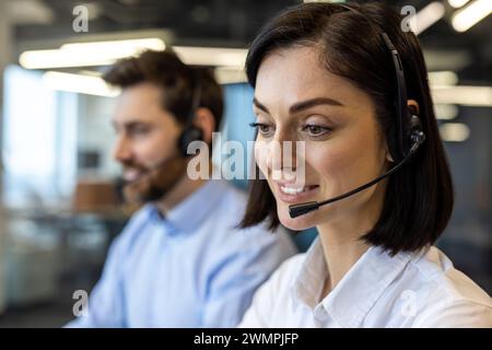 Portrait of smiling young woman in wireless headset and white shirt looking ahead with smile next to male coworker. Call centre employees starting new day and ready for providing consulting services. Stock Photo