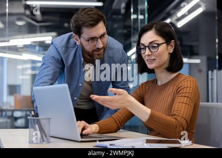 Confident caucasian secretary in eyeglasses showing screen of laptop to attentive bearded head of company. Successful coworkers discussing plans and goals for week while making list of meetings. Stock Photo