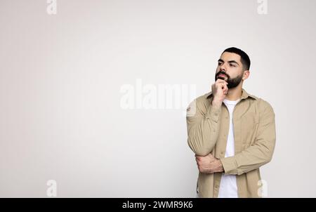 Pensive man with a beard in a beige jacket and white shirt holds chin, contemplating Stock Photo