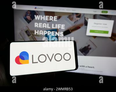 Person holding mobile phone with logo of German online dating platform company LOVOO in front of business web page. Focus on phone display. Stock Photo
