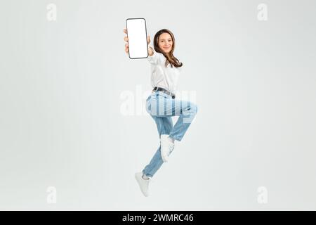 Dynamic young woman in mid-air jump, joyfully presenting a smartphone with a blank screen Stock Photo