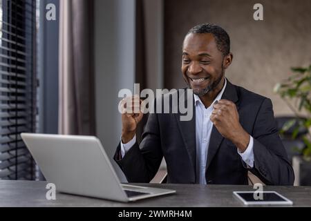 Ecstatic African American businessman fist pumping in triumph at his workplace with a laptop and tablet. Stock Photo