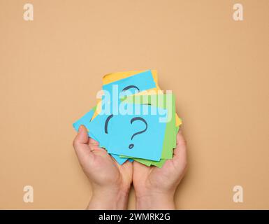 Female hands hold paper sheets with a drawn question mark on a beige background, searching for the answer and truth Stock Photo